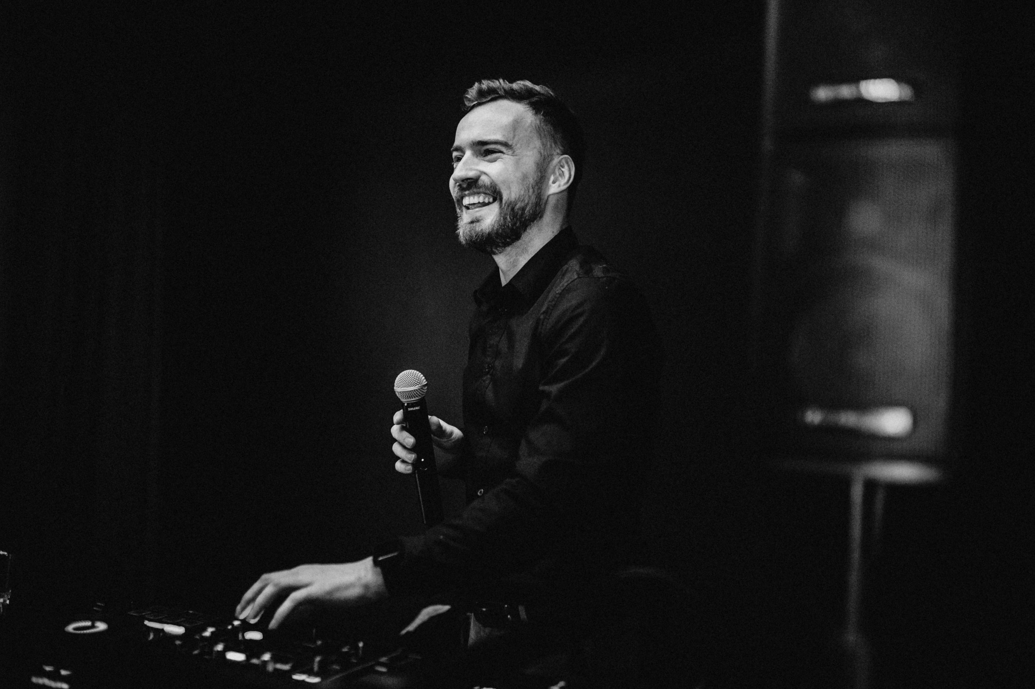 Uśmiechnięty DJ w czarnej koszuli, trzymający mikrofon Shure, miksuje muzykę na konsolecie w ciemnym otoczeniu.