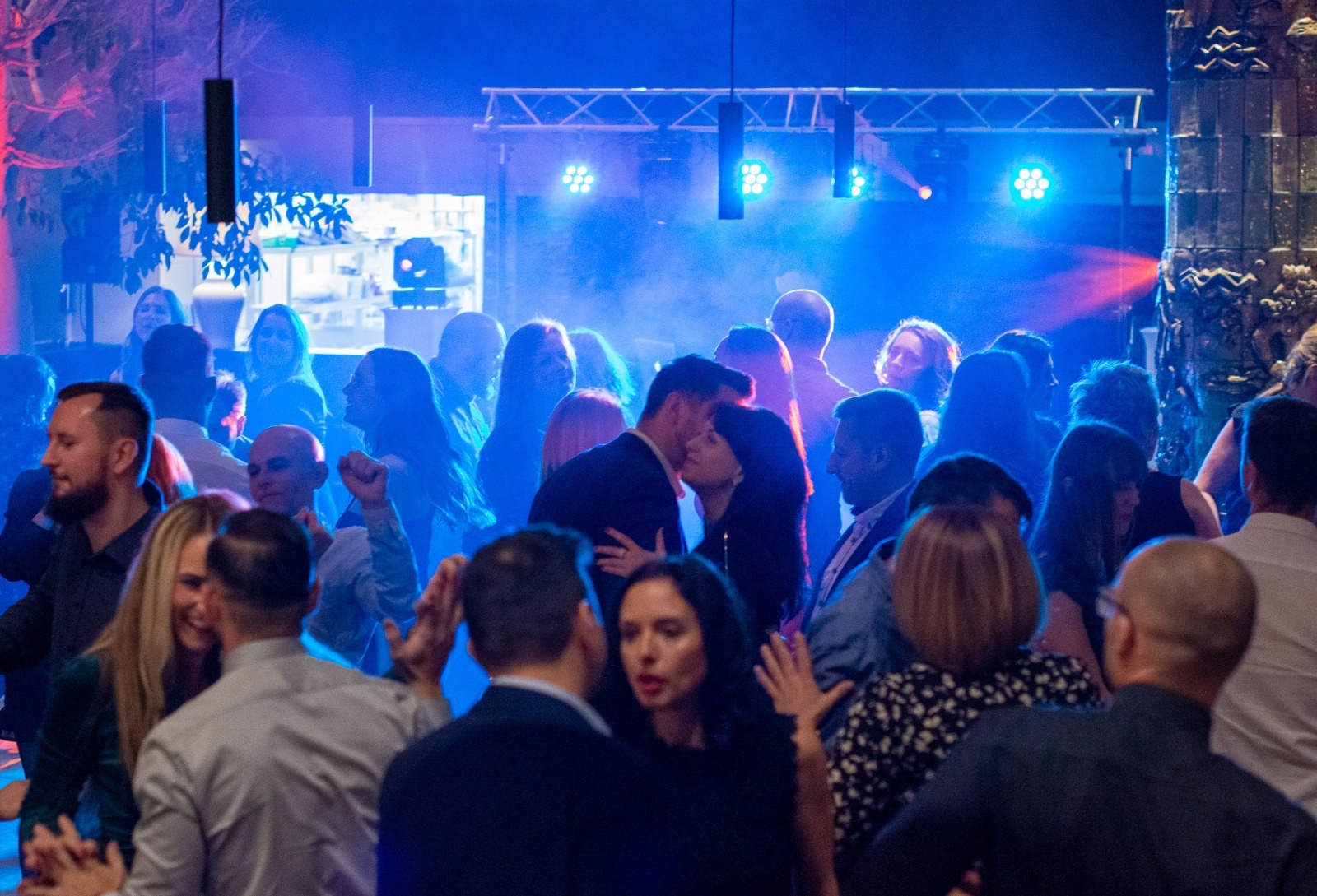 Para całująca się na parkiecie podczas imprezy oświetlonej niebieskim światłem, widoczni inni uczestnicy zabawy i elementy scenografii.