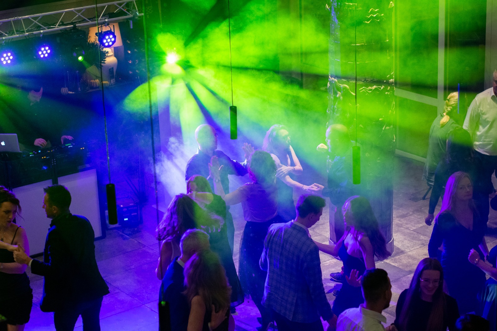 Roztańczony tłum ludzi w klubie oświetlonym zielonymi i niebieskimi światłami, DJ grający muzykę w tle za parawanem.