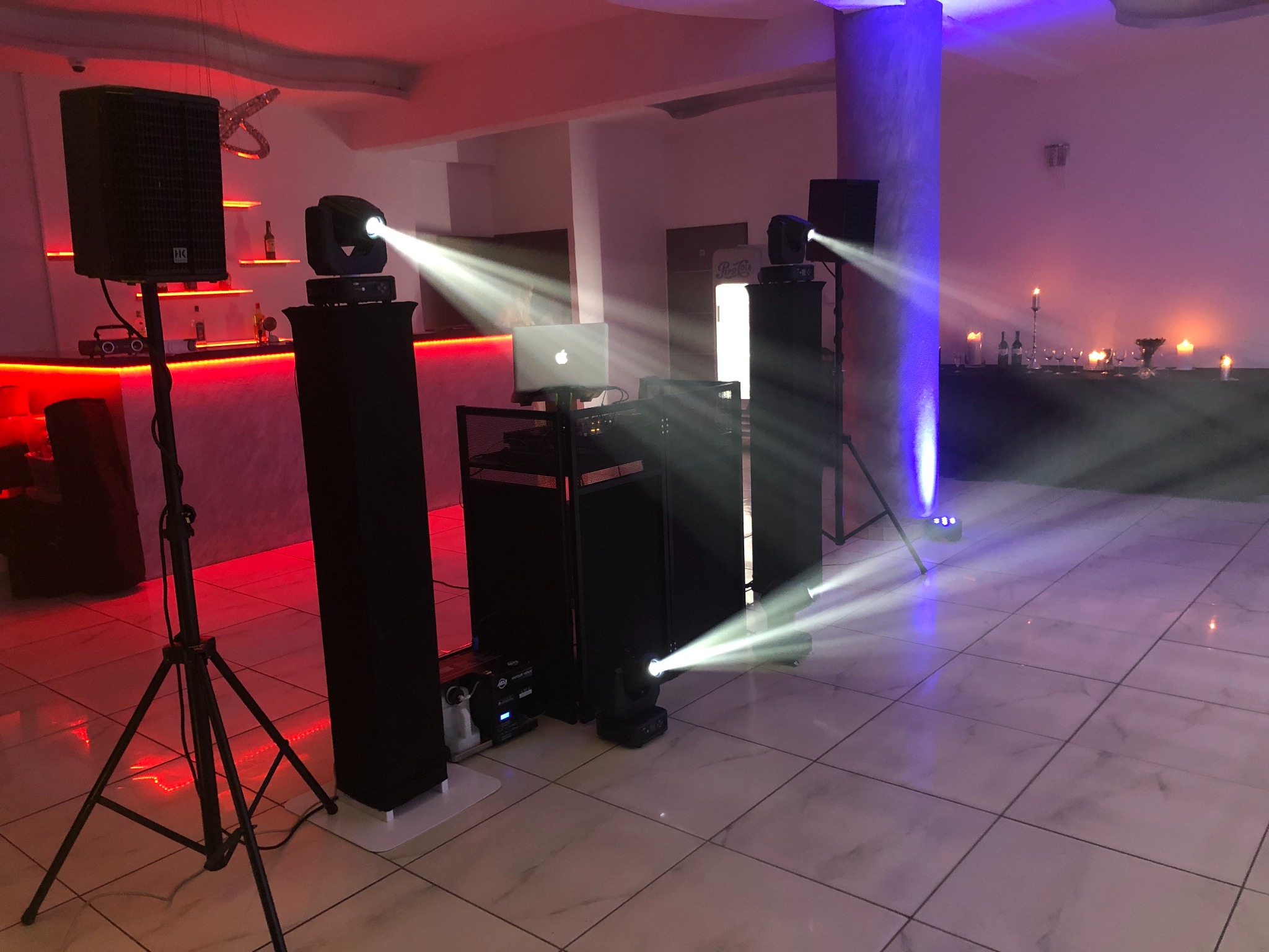 Sala bankietowa oświetlona reflektorami scenicznymi i nagłośniona głośnikiem na statywie, bar z podświetleniem LED w tle, laptop na stanowisku DJ-a.