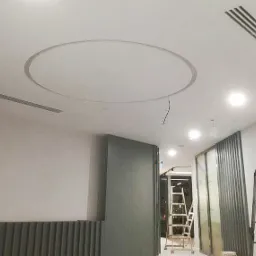 Biuro po remoncie: Biały sufit z okrągłym wgłębieniem i kratkami wentylacyjnymi, widoczne fragmenty ścian i drabina w tle.