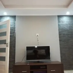 Telewizor na szafce RTV na tle szarej ściany z dekoracyjnym kamieniem i wystającym kablem zasilającym.