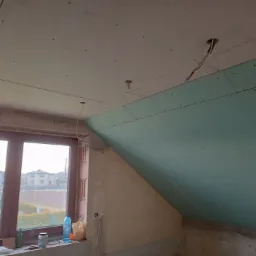 Poddasze w trakcie montażu płyt karton gips, widoczne szpachlowane łączenia płyt na suficie i skośnej ścianie, wystające kable oświetleniowe, okno z widokiem na domy.