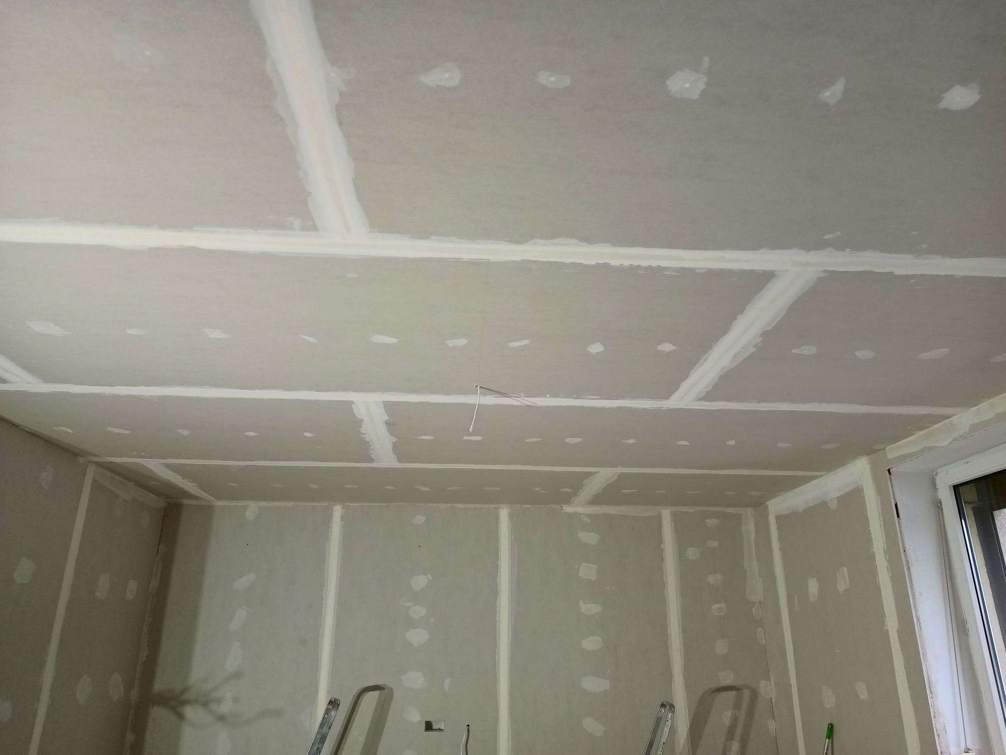 Montaż płyt karton gips na ścianach i suficie, widoczne łączenia szpachlowane, przewód elektryczny wystający z sufitu, drabina aluminiowa w rogu pomieszczenia.