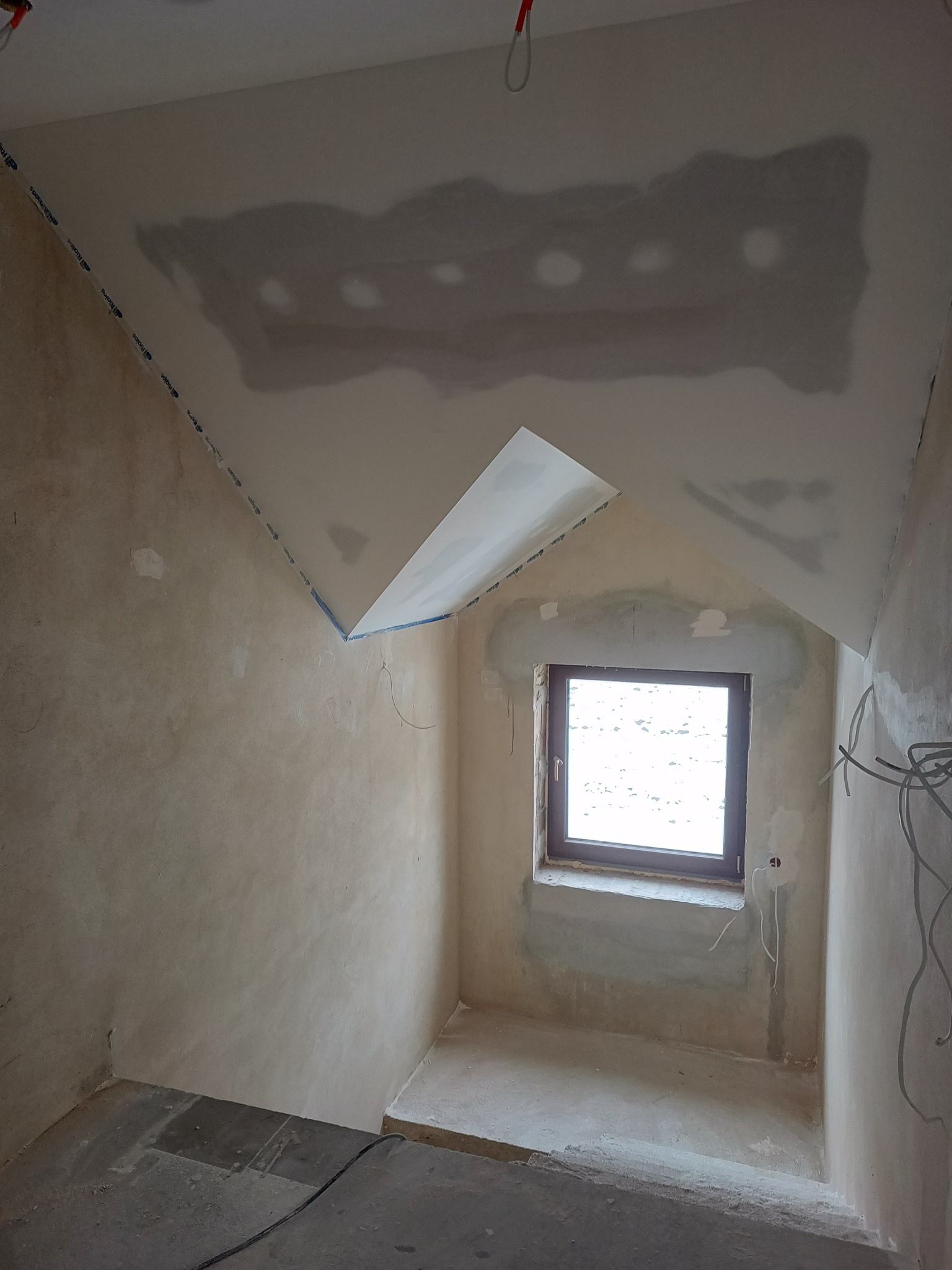Pokój na poddaszu w trakcie remontu: nierówna ściana z widocznymi śladami po wcześniejszych pracach, częściowo wykończony sufit z taśmą malarską przy krawędziach oraz widoczne otwory po...