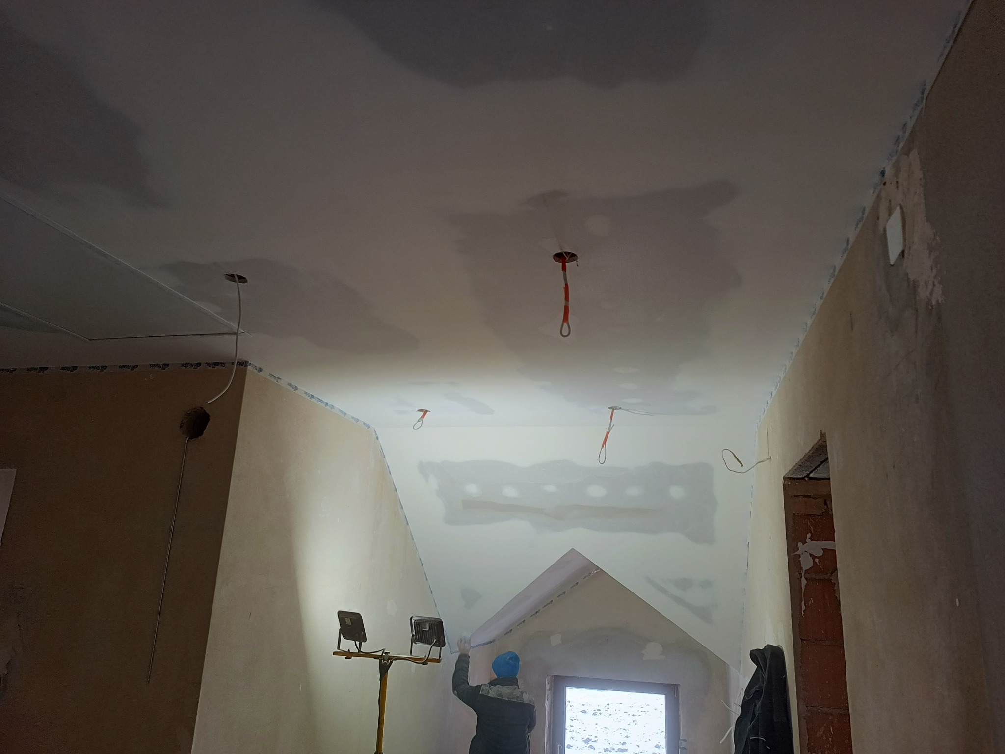 Prace wykończeniowe w pokoju na poddaszu: mężczyzna w niebieskiej czapce szpachluje sufit z widocznymi przewodami oświetleniowymi, oświetlany przez lampy budowlane.