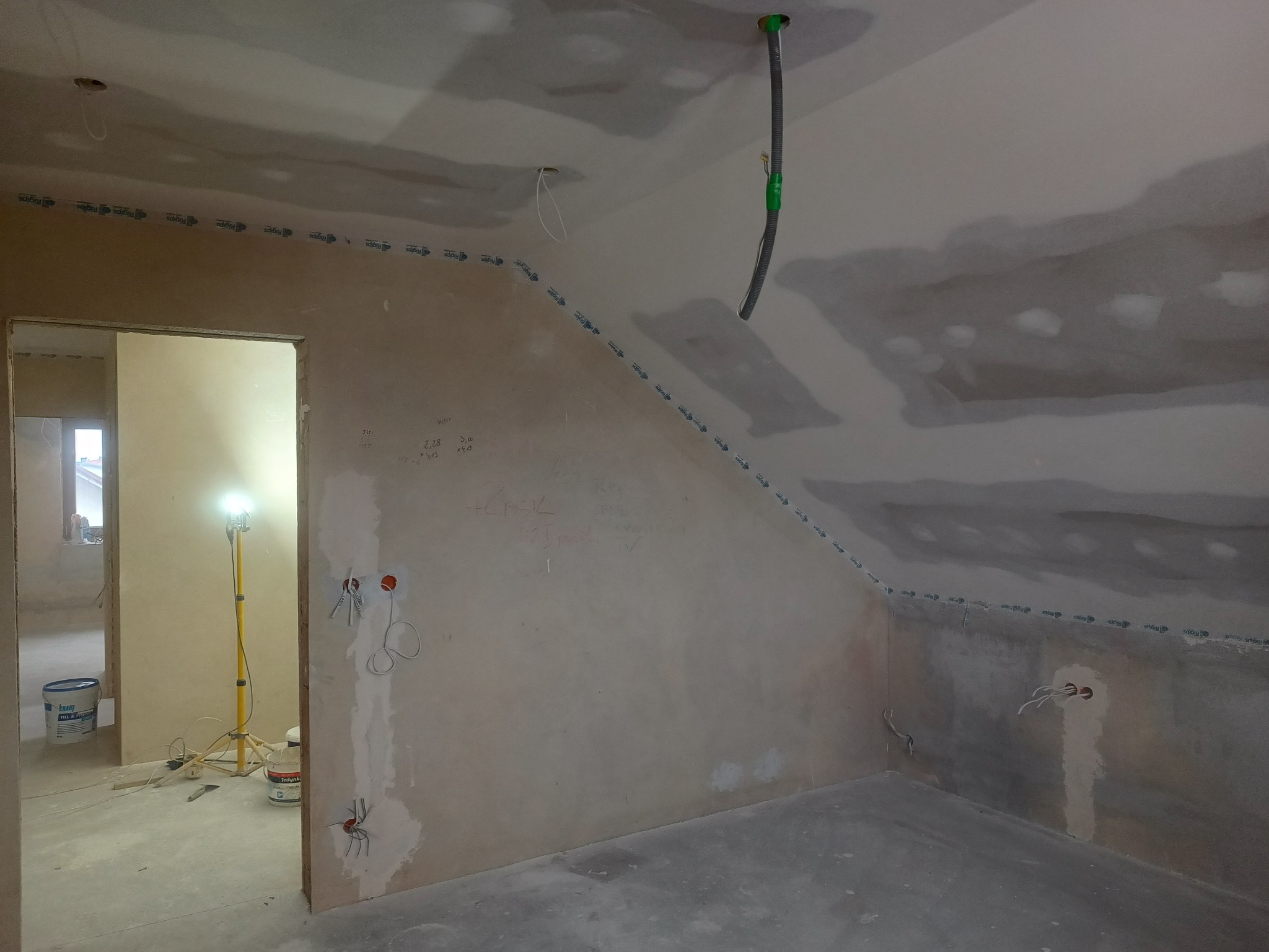 Pokój na poddaszu w trakcie remontu: ściany przygotowane do malowania, z widocznymi przewodami elektrycznymi, taśmą Rigips i śladami po szpachlowaniu, z otwartym przejściem do kolejnego...