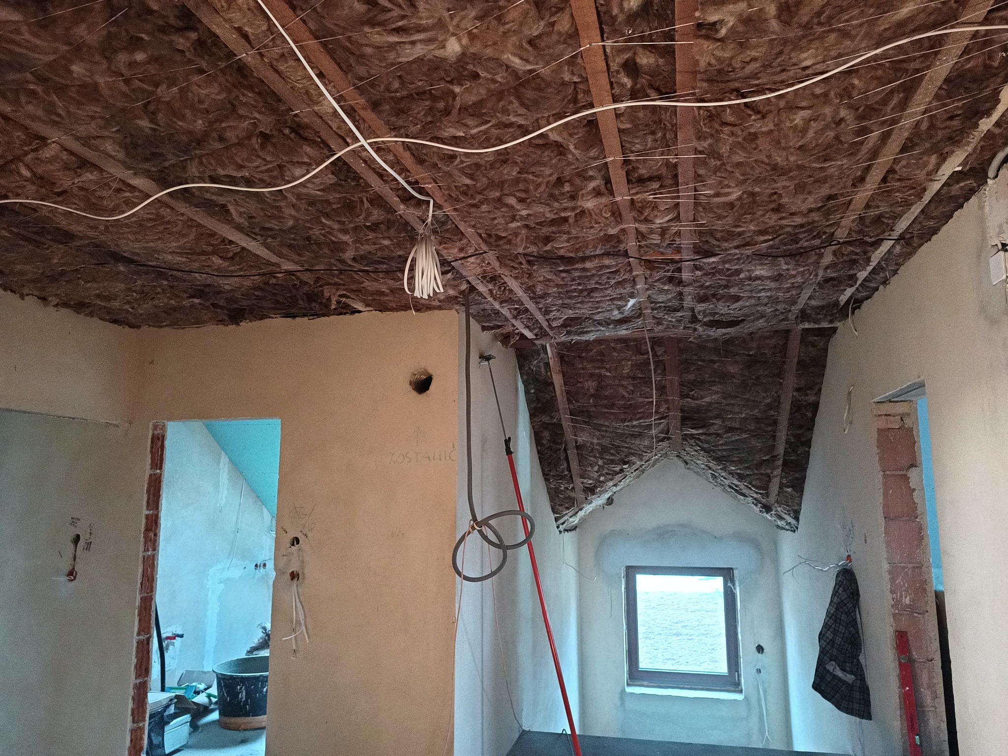 Poddasze w trakcie remontu: izolacja wełną mineralną widoczna na suficie, przewody elektryczne, ściany przygotowane do dalszych prac wykończeniowych, okno z widokiem na morze.