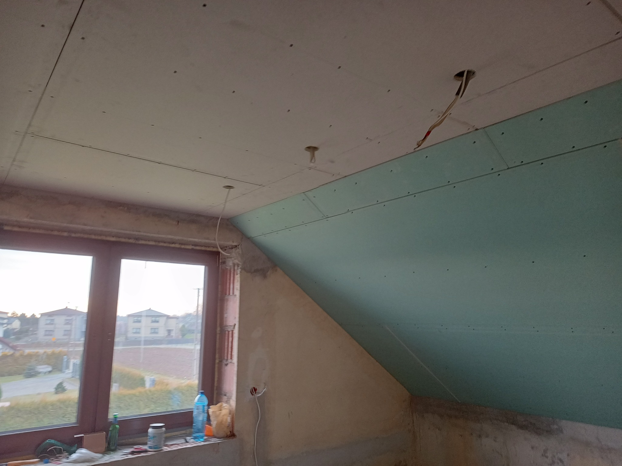 Poddasze w trakcie montażu płyt karton gips, widoczne szpachlowane łączenia płyt na suficie i skośnej ścianie, wystające kable oświetleniowe, okno z widokiem na domy.