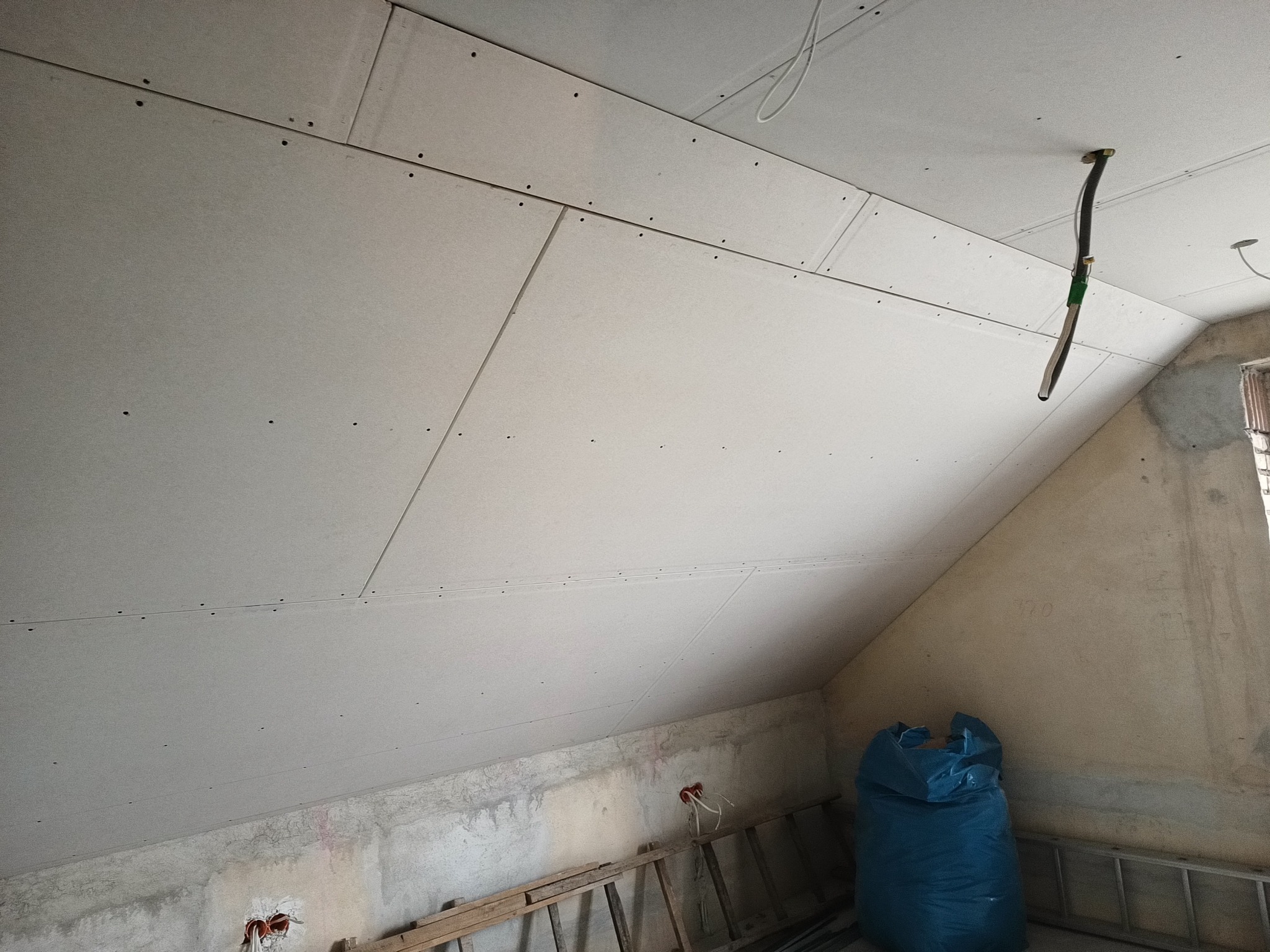 Skośny sufit na poddaszu w trakcie montażu, widoczne płyty z przykręconymi wkrętami, wystające kable i nieotynkowane ściany z instalacją elektryczną oraz drabina i worek z materiałami.