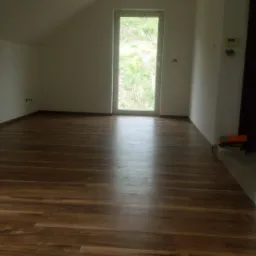 Pokój na poddaszu w Warszawie z nową, ciemną podłogą z paneli, widok na okno i fragment ściany z wystającymi kablami.