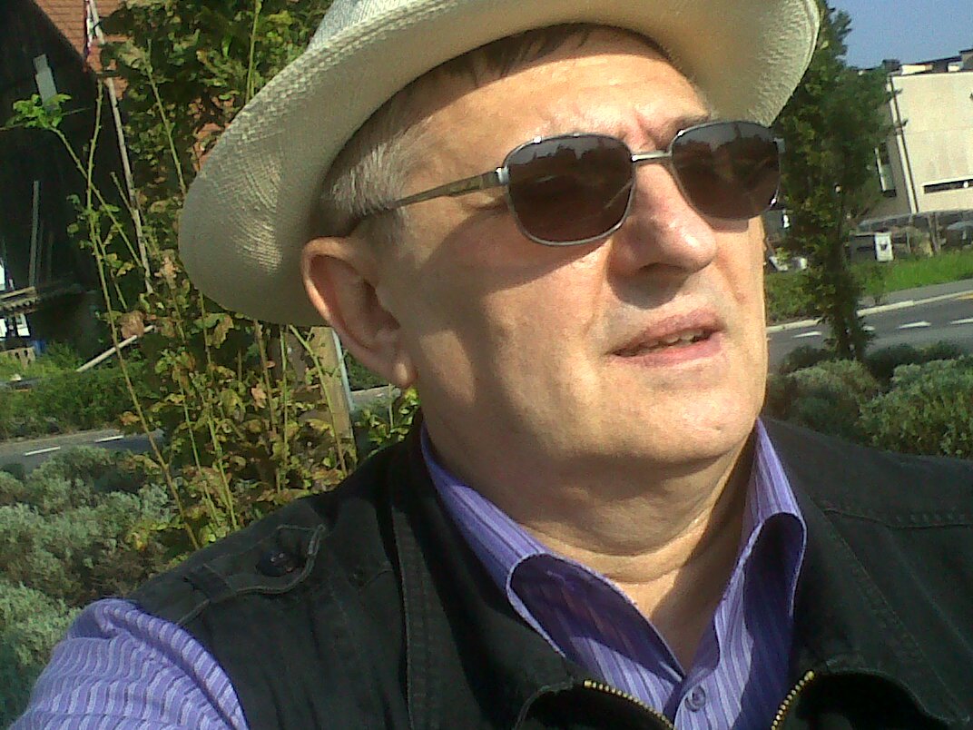 Portret mężczyzny w kapeluszu i okularach przeciwsłonecznych, w tle zabudowania i zieleń.