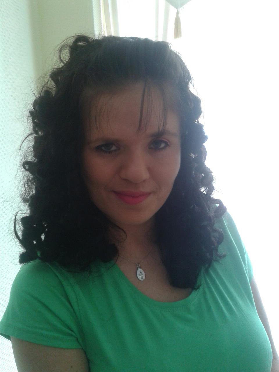 Portret kobiety z kręconymi, ciemnymi włosami, ubranej w zieloną koszulkę, z delikatnym srebrnym naszyjnikiem na szyi, na tle jasnej ściany i okna.