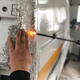 Blacha samochodu dostawczego z widocznymi śladami naprawy, przytrzymywana dłonią, podczas nagrzewania punktowego za pomocą specjalistycznego urządzenia.