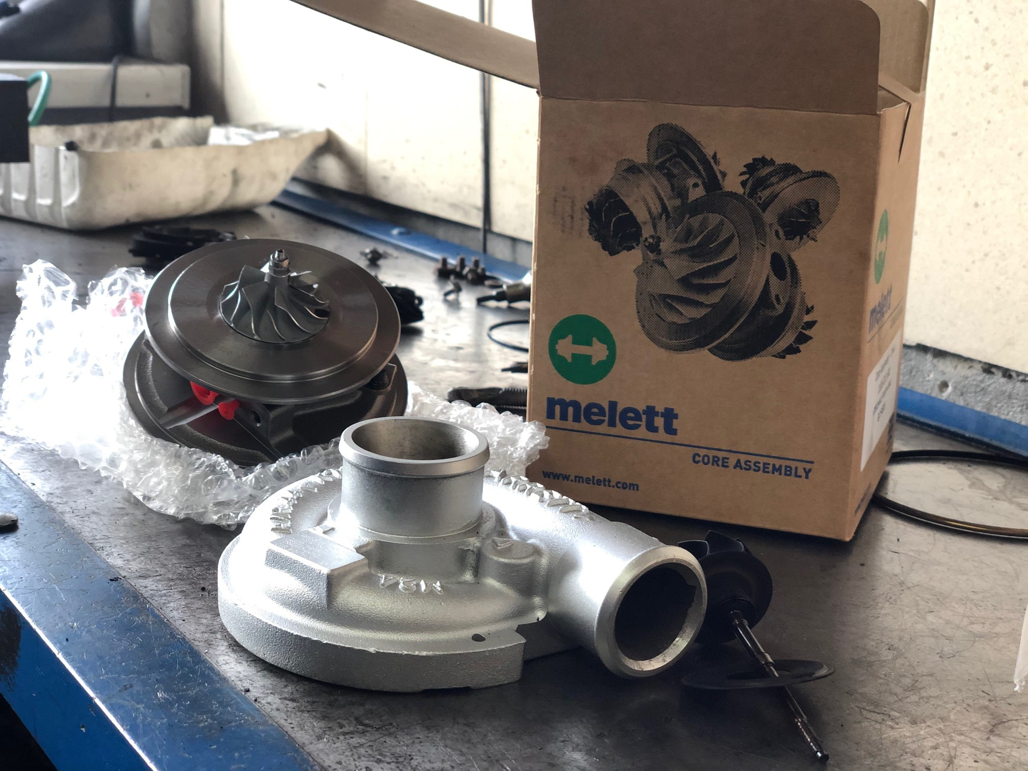 Nowa turbosprężarka samochodowa marki Melett, rozpakowana z pudełka, leży na warsztatowym stole.