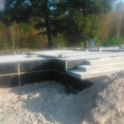 Zabezpieczone fundamenty domu jednorodzinnego z izolacją pionową i schodami wejściowymi, otoczone piaskiem i zielenią drzew w tle.