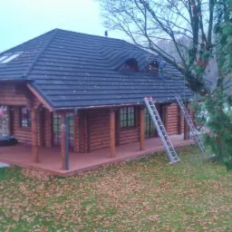 Drewniany dom z ciemnym dachem karpiówką i dwoma oknami dachowymi, z werandą, na której stoją dwie drabiny. Jesienne liście na trawniku.