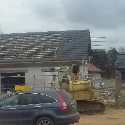 Budowa domu jednorodzinnego: widok na częściowo pokryty dach z łatami, z surowymi ścianami i żółtą koparką na działce, szary samochód na pierwszym planie.