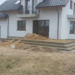 Nowo wybudowany dom jednorodzinny z betonu komórkowego z widocznymi schodami wejściowymi w trakcie budowy, pracownik w szarym dresie stoi obok.