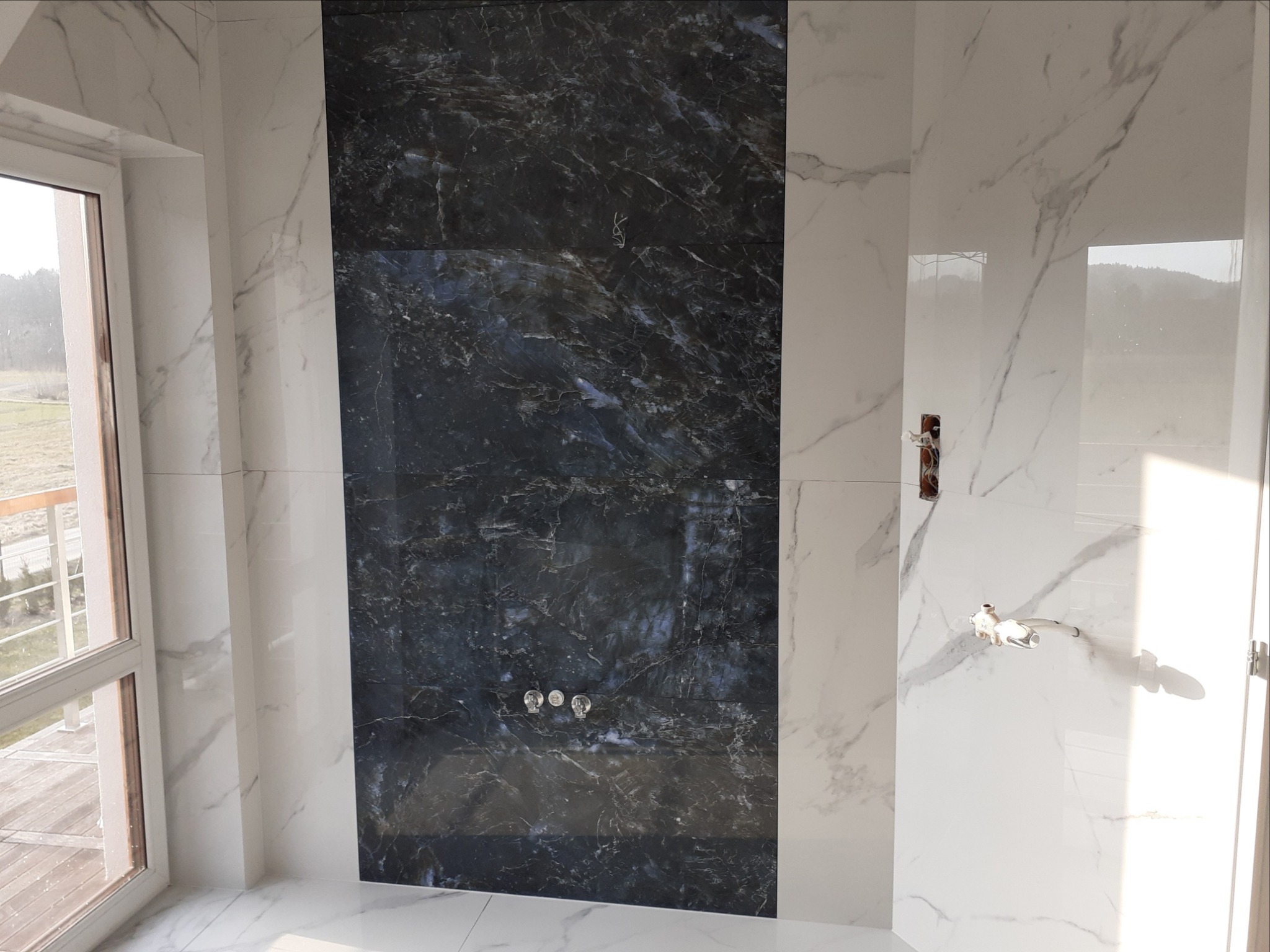 Ściana łazienki wyłożona białymi i czarnymi płytkami imitującymi marmur, z widocznymi przyłączami do armatury i oknem z widokiem na zewnątrz.