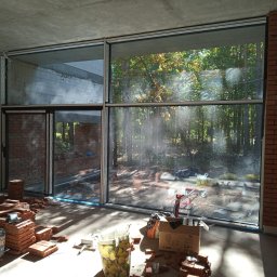 M-Stal Bartosz Mazik - Duże okna aluminiowe z widokiem na las w surowym wnętrzu z ceglanymi ścianami i stertami cegieł. W tle widoczny wózek transportowy.