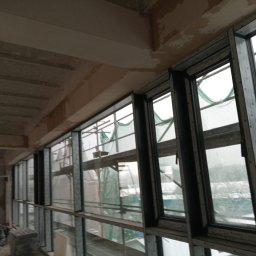 M-Stal Bartosz Mazik - Rząd okien aluminiowych z widocznymi rusztowaniami na zewnątrz, zamontowanych w surowym stanie budynku. Widok z wnętrza, szare, pochmurne tło za oknami.