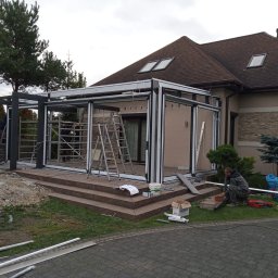 M-Stal Bartosz Mazik - Montaż przeszklonej konstrukcji tarasowej przy domu jednorodzinnym. Widoczne elementy konstrukcyjne, drabiny i pracownicy podczas instalacji. Etap budowy.