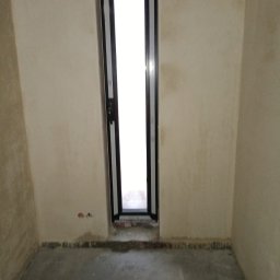 M-Stal Bartosz Mazik - Wąskie, czarne okno aluminiowe w surowym wnętrzu. Widoczne ślady po montażu na ścianie i podłodze. Minimalistyczny design ramy okiennej. Jasne światło wpada przez szybę.