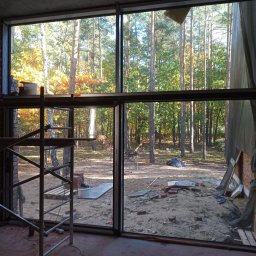 M-Stal Bartosz Mazik - Montaż okna aluminiowego z widokiem na las jesienią. Widoczne rusztowanie i murarz. Duże przeszklenie w surowym stanie budynku.