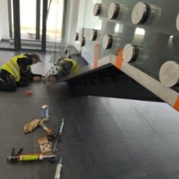 M-Stal Bartosz Mazik - Montaż szklanej balustrady z użyciem mocowań punktowych. Dwóch monterów w kamizelkach odblaskowych przy pracy, narzędzia na podłodze. Widok z boku, wnętrze budynku.