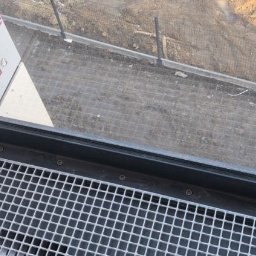 M-Stal Bartosz Mazik - Metalowa krata pod oknem, widok z góry, z elementami mocującymi. W tle widoczny teren budowy z siatką i ziemią. Ujęcie z bliska, ukazujące detale konstrukcji.