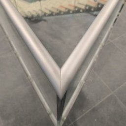 M-Stal Bartosz Mazik - Detal narożnika balustrady szklanej z aluminiowym profilem, widok z góry na szare płytki podłogowe i schody w tle. Precyzyjne łączenie profili.