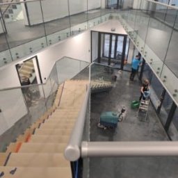 M-Stal Bartosz Mazik - Wnętrze budynku z przeszklonymi balustradami i schodami zabezpieczonymi kartonem. Osoby czyszczą posadzkę przy użyciu maszyny i drabiny. Widok z góry.