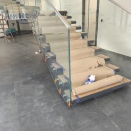 M-Stal Bartosz Mazik - Nowoczesne schody z balustradą ze szkła, zabezpieczone kartonem na stopniach. Widoczne metalowe elementy konstrukcyjne i szklane panele. Minimalistyczny design.