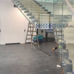 M-Stal Bartosz Mazik - Nowoczesne schody z balustradą ze szkła hartowanego, mocowane punktowo. Widoczna drabina i maszyna czyszcząca, co sugeruje etap końcowy prac montażowych.