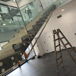 M-Stal Bartosz Mazik - Szklane schody z metalowymi mocowaniami, widoczne z dołu, z drabiną w tle. Minimalistyczny design, nowoczesne wykończenie wnętrza, czarna podłoga.