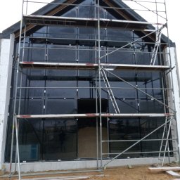 Montaż okien w budynku o nowoczesnej architekturze. Widoczne rusztowania i duże przeszklenia. Budynek w trakcie wykończenia, surowy stan.