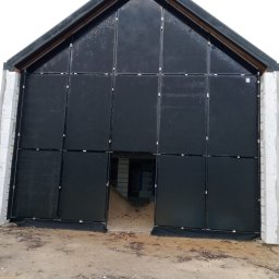 M-Stal Bartosz Mazik - Ciemna fasada budynku w trakcie budowy, podzielona na panele z widocznymi mocowaniami. Surowy stan, widoczne bloczki i ziemia. Ujęcie z dołu, podkreślające wysokość.