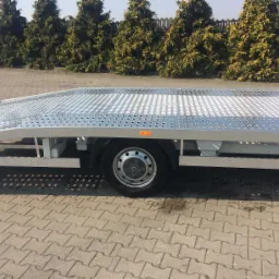 Galwanizowana platforma niskopodwoziowa przyczepy samochodowej z perforowaną powierzchnią, widoczne koło i elementy ramy, na tle brukowana nawierzchnia i rząd tuj.