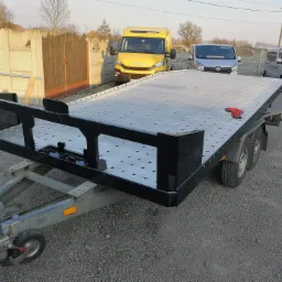 Nowa, metalowa przyczepa samochodowa z platformą z otworami, pomalowana na szaro i czarno, stojąca na żwirowym placu, z widocznymi samochodami w tle i czerwoną rękawicą roboczą na platformie.