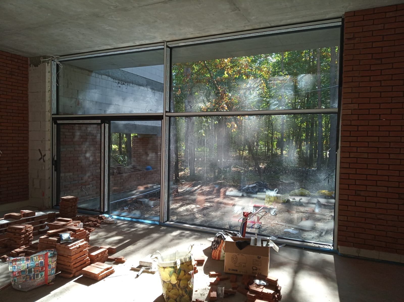 Duże okna aluminiowe z widokiem na las w surowym wnętrzu z ceglanymi ścianami i stertami cegieł. W tle widoczny wózek transportowy.