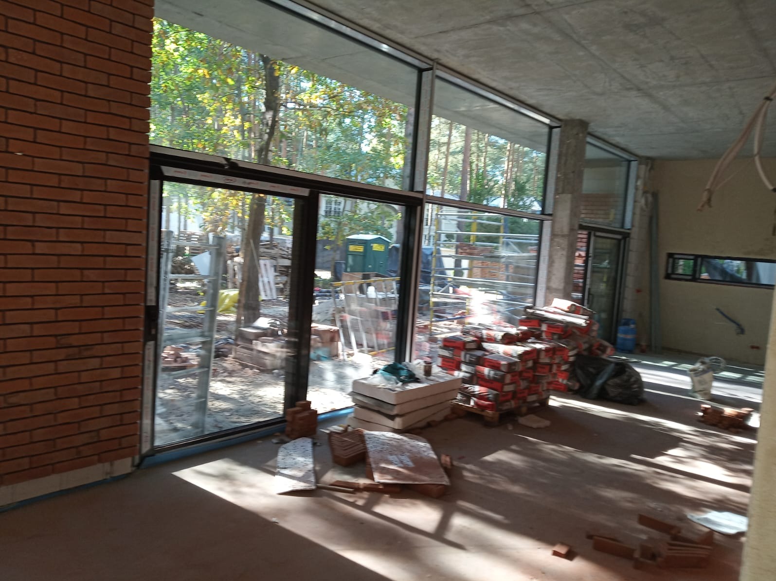Wnętrze w trakcie budowy z dużymi oknami aluminiowymi i stertą materiałów budowlanych. Widok na zewnątrz z drzewami. Surowy betonowy sufit i ceglana ściana.