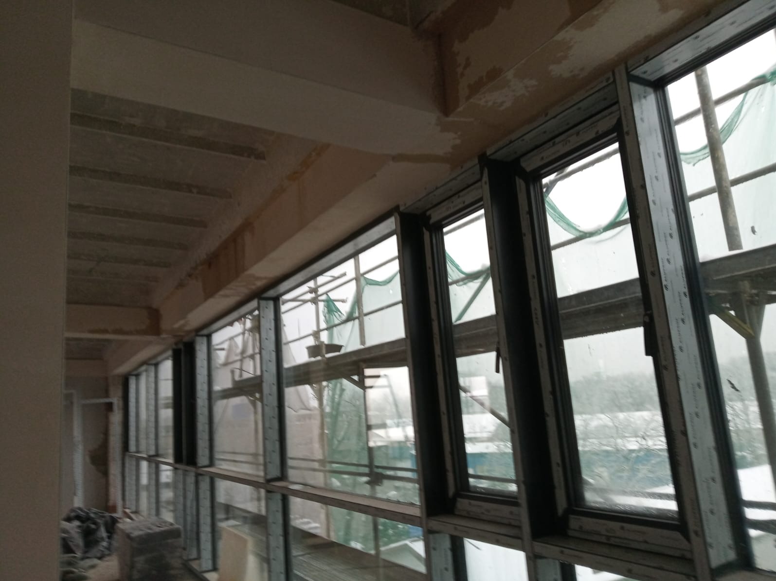 Rząd okien aluminiowych z widocznymi rusztowaniami na zewnątrz, zamontowanych w surowym stanie budynku. Widok z wnętrza, szare, pochmurne tło za oknami.