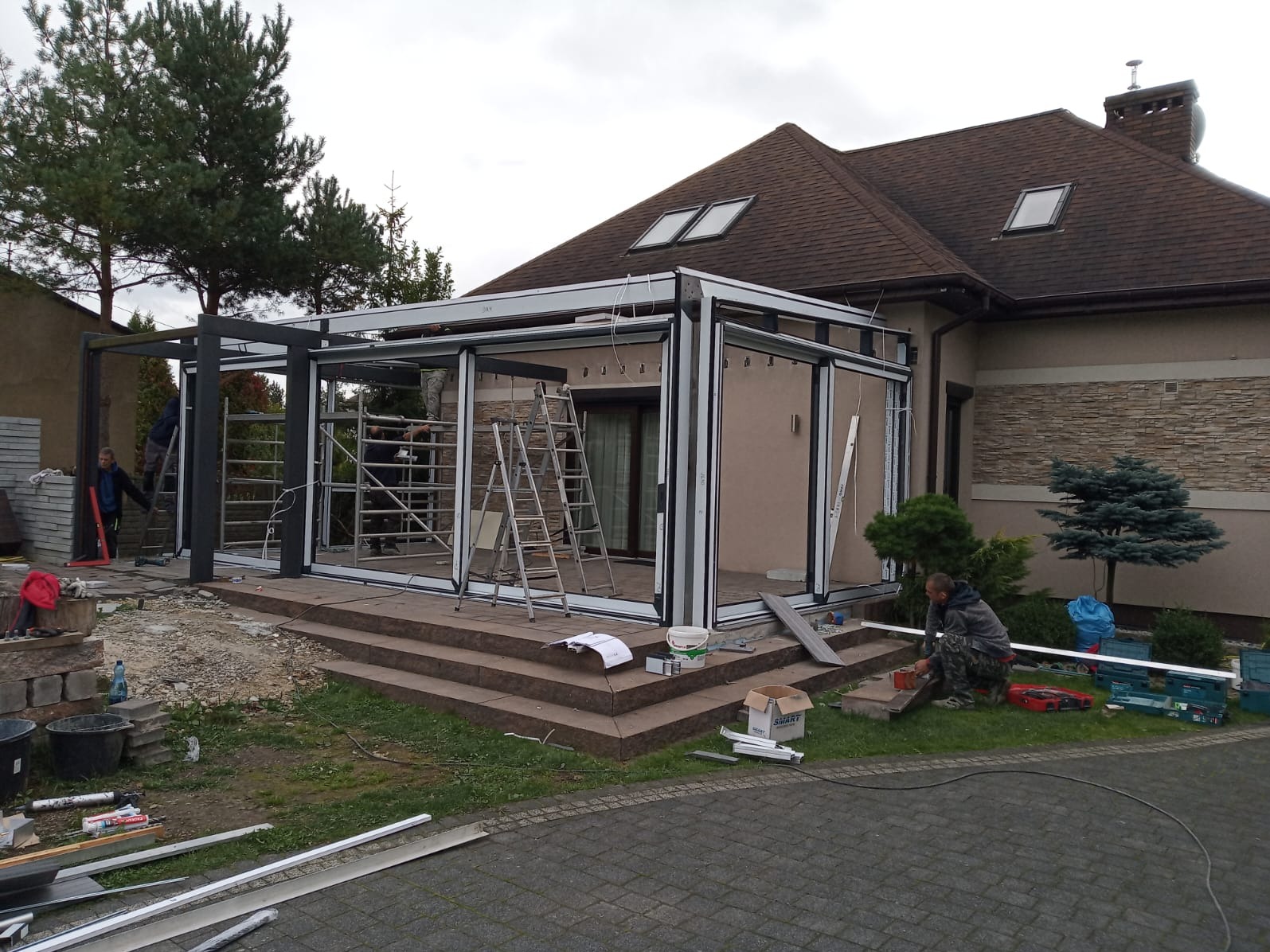 Montaż przeszklonej konstrukcji tarasowej przy domu jednorodzinnym. Widoczne elementy konstrukcyjne, drabiny i pracownicy podczas instalacji. Etap budowy.