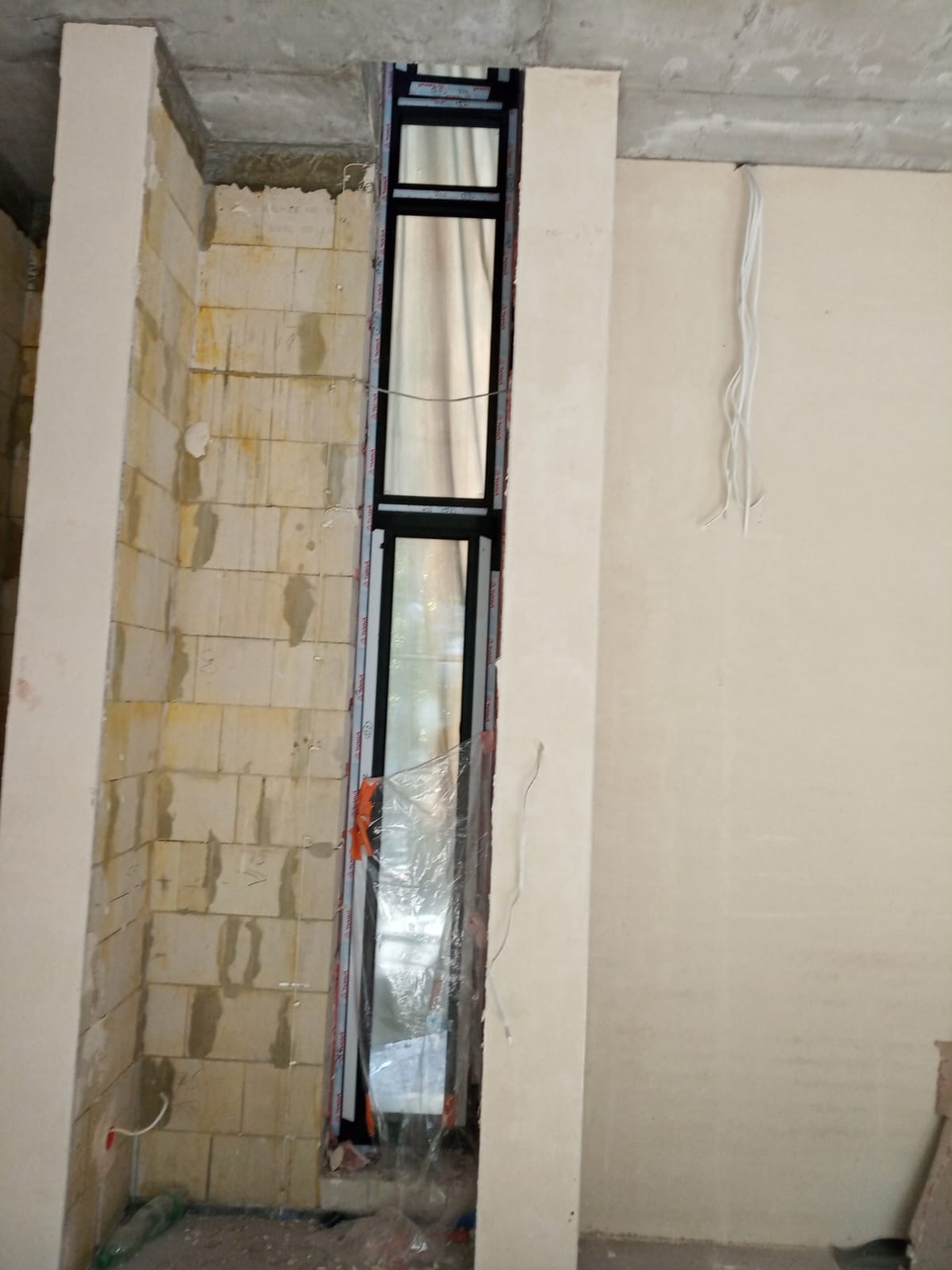 Wąskie okno PCV z czarną ramą, w trakcie montażu w surowym otworze ściennym z pustaków. Widoczne kable elektryczne i folia ochronna na oknie.
