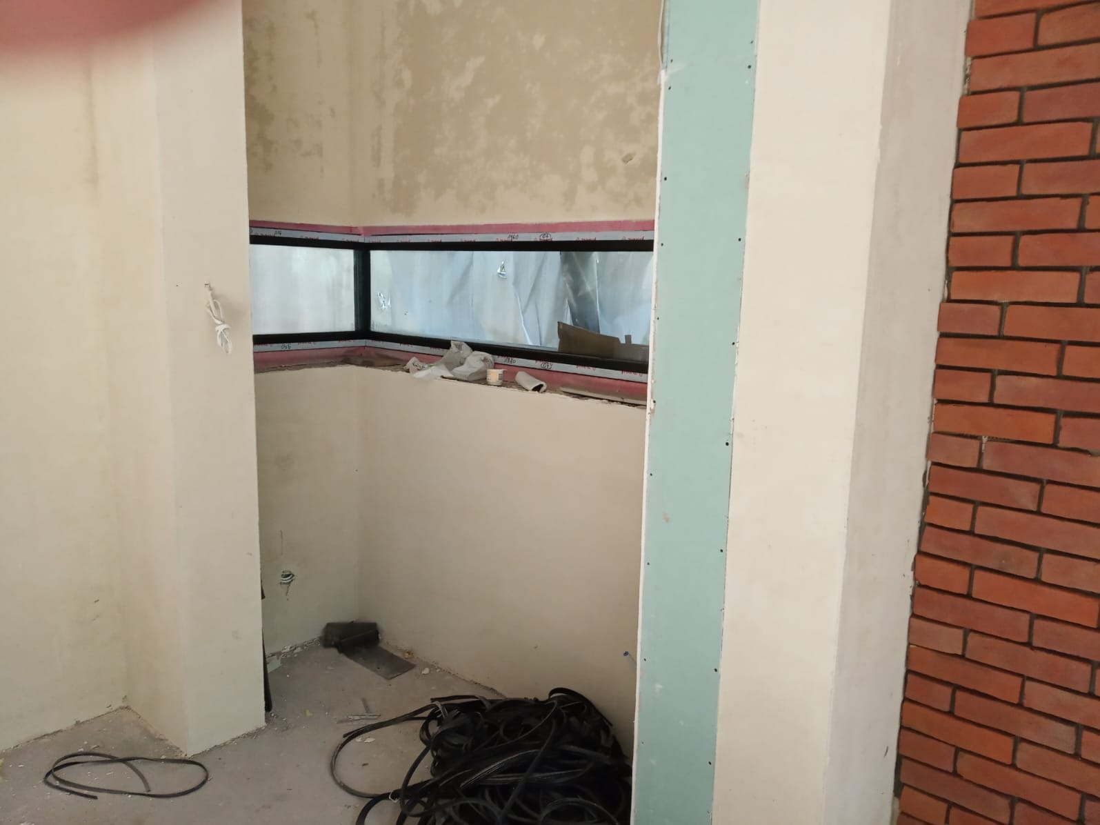 Wnętrze w trakcie wykończenia: nietypowe okno z czarną ramą, fragment ściany z cegły, przewody na podłodze. Widoczne płyty gipsowo-kartonowe.
