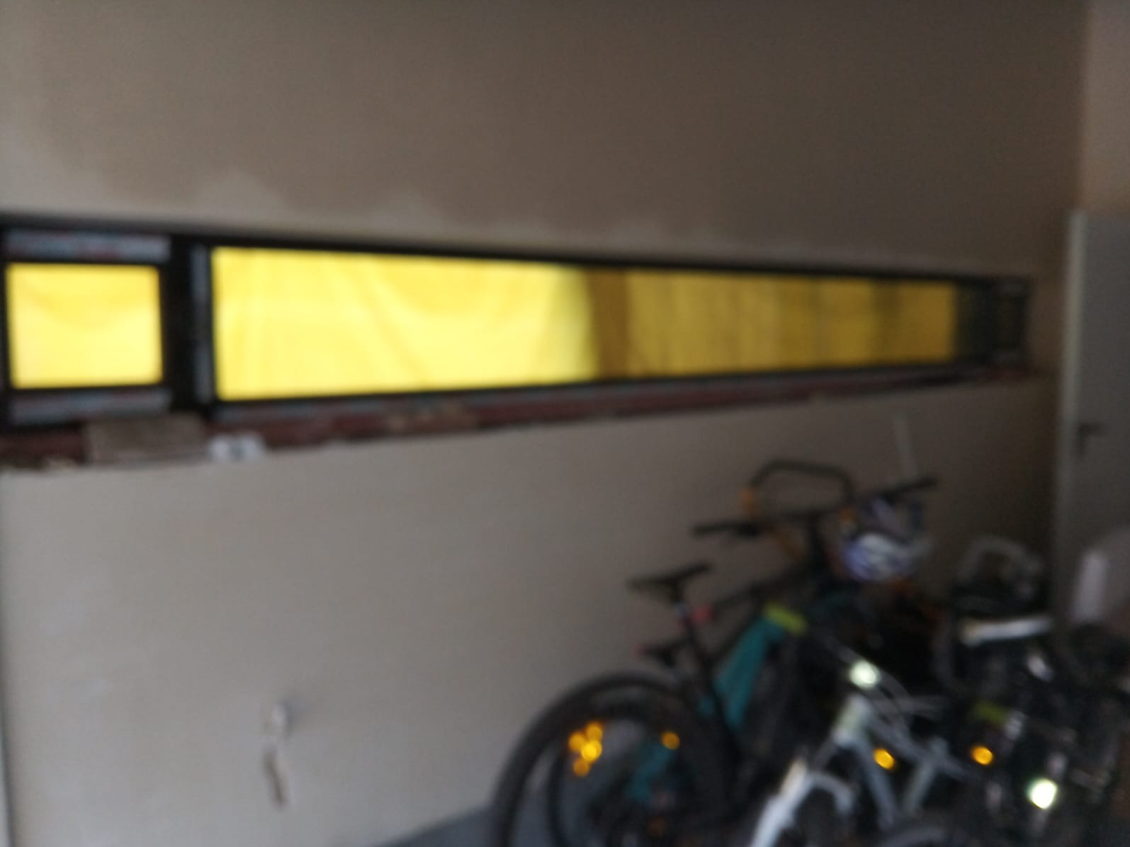 Długie, wąskie okno z czarną ramą, w trakcie montażu. Widoczne fragmenty muru i żółte światło wpadające do wnętrza. W tle rowery, co sugeruje garaż lub piwnicę.