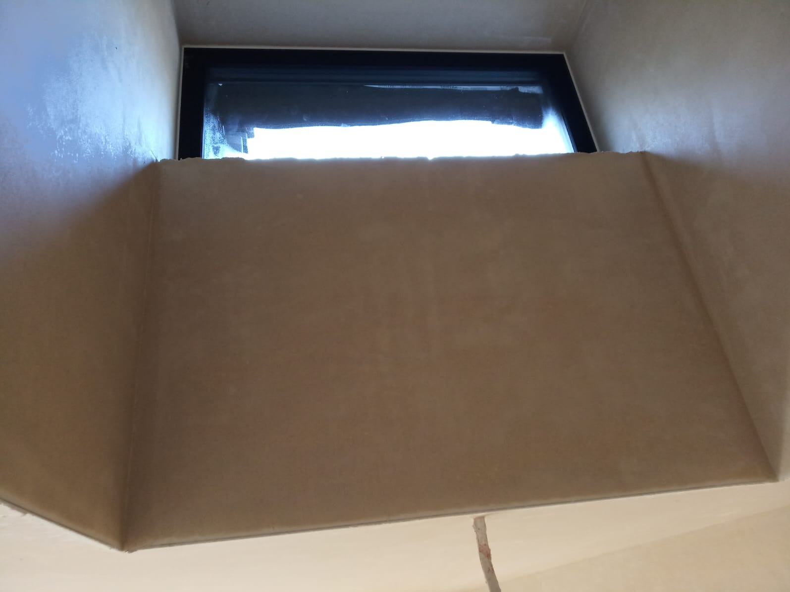 Zamontowane okno w ścianie z wykończonym wnęką, widok od dołu. Ściana w odcieniach beżu, okno z czarną ramą. Widoczne światło dzienne wpadające przez szybę.