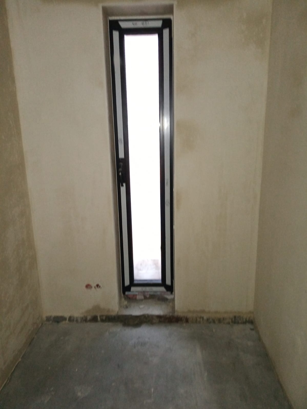 Wąskie, czarne okno aluminiowe w surowym wnętrzu. Widoczne ślady po montażu na ścianie i podłodze. Minimalistyczny design ramy okiennej. Jasne światło wpada przez szybę.