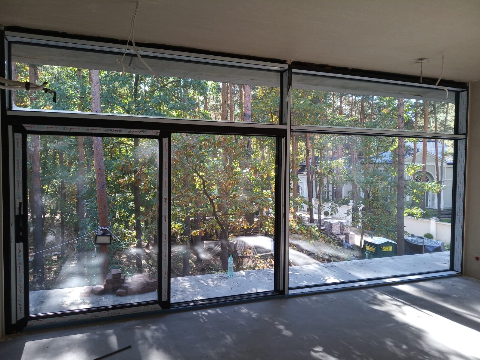 Duże, przesuwne okna aluminiowe z czarnymi ramami, widok na zalesiony teren i dom. Surowy stan wykończenia wnętrza, betonowa podłoga, widoczne przewody.